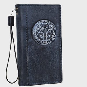 Könnte beinhalten: Eine dunkelblaue Lederbrieftasche mit einem kreisförmigen, geprägten Emblem einer Kobra auf der Vorderseite. Die Brieftasche verfügt über eine passende dunkelblaue Handschlaufe, die mit einem kleinen Metallring und einem schwarzen Schiebeverschluss befestigt ist.