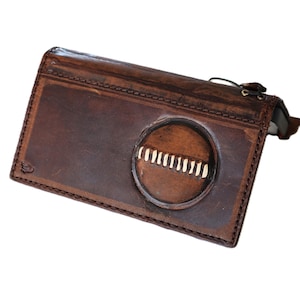 May include: A brown leather wallet with a circular design on the front. The wallet has a rectangular shape with a stitched border and a small, decorative emblem on the lower left corner. The circular design has a series of vertical white lines.