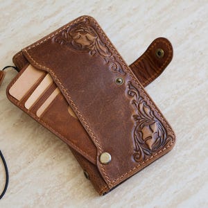 May include: A brown leather wallet case featuring intricate floral tooling and embossed patterns. The wallet includes multiple interior card slots, a snap button closure, and a thin black wrist strap attached to the side for carrying.