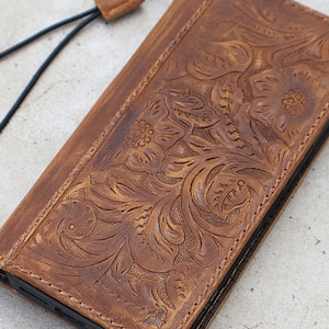 Könnte beinhalten: Eine braune Leder-Handyhülle mit floralem Prägemuster. Die Hülle hat einen genähten Rand und eine schwarze Kordel oben. Das Leder hat ein Used-Aussehen.