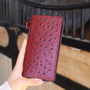 Peut inclure: Un portefeuille en cuir bordeaux avec un motif autruche texturé. Le portefeuille rectangulaire a une petite boucle sur le côté. Les coutures sont rouges.