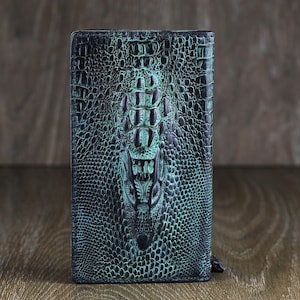 Puede incluir: Una cartera de cuero azul oscuro con un patrón de piel de cocodrilo y un acento verde azulado.