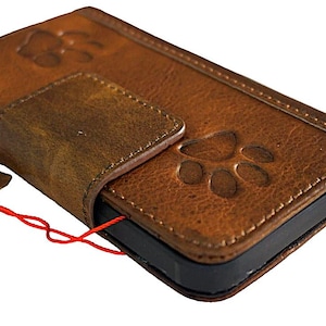 Puede incluir: Una funda de teléfono de cuero marrón con cierre de solapa y diseños de huellas de patas. La funda tiene un aspecto desgastado y está hecha de cuero genuino.