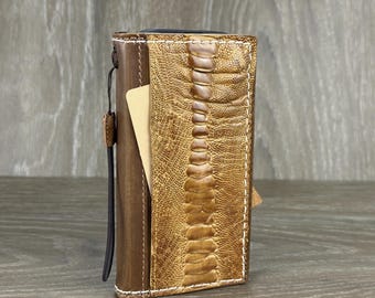 Könnte beinhalten: Ein braunes Leder-Handy-Etui mit einem hellbraunen Straußenledermuster. Das Etui hat eine Klappe, die mit einem Druckknopf geschlossen wird, und einen Riemen, der zum Tragen des Telefons verwendet werden kann.