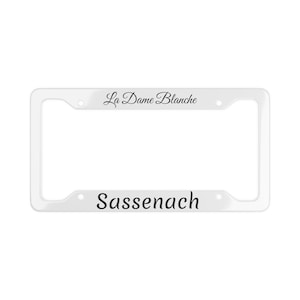 Puede incluir: Marco de matrícula de plástico blanco con el texto "La Dame Blanche" y "Sassenach" en fuente cursiva negra.