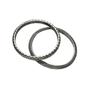 May include: Two silver-colored bangles with a textured, woven pattern. The bracelets are circular and overlap slightly, set against a white background. The design features a repeating square pattern, giving a geometric look.