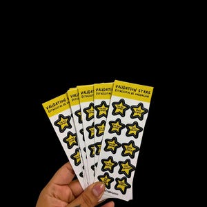 May include: A set of five vertical sticker sheets featuring yellow star-shaped stickers with black outlines. Each star contains text in English and Spanish, such as You Are Capable, Eres Capaz, You Are Loved, and Eres Querida. The header reads Validation Stars.
