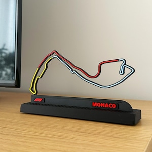Peut inclure: Une base noire avec un motif en fibre de carbone et le texte "MONACO" en rouge. Un modèle 3D du circuit de Formule 1 de Monaco est placé sur la base. La piste est en plastique rouge, jaune et bleu.