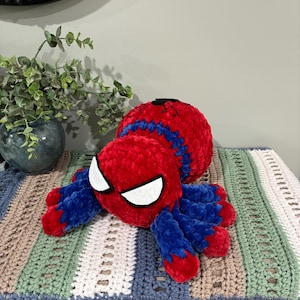 May include: A handmade plush Spider-Man toy in red and blue. The plush features a round head with white eye details and eight blue and red legs. The toy is made of a soft, textured material.