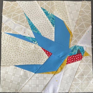 May include: A square fabric block featuring a blue bird design. The bird is constructed from various fabric pieces in shades of blue, white, beige, and red. The bird's body is a solid blue, with wings and tail feathers in patterned fabrics.