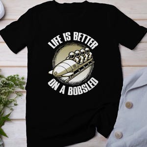 Puede incluir: Camiseta negra con un gráfico blanco de cuatro personas en un trineo. El gráfico está rodeado de un círculo con el texto "Life is better on a bobsled".