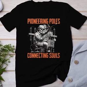 May include: Black t-shirt with a graphic of a skeleton working on a power pole. The text "Pioneering Poles Connecting Souls" is printed on the shirt.