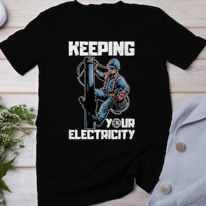 Op de afbeelding: Zwart T-shirt met een afbeelding van een elektricien die een elektriciteitspaal beklimt. De tekst "KEEPING YOUR ELECTRICITY" staat op het shirt gedrukt.