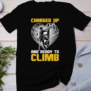 May include: Black t-shirt with a yellow graphic of a lineman climbing a utility pole inside a heart shape. The text "CHARGED UP" is above the graphic and "AND READY TO CLIMB" is below.