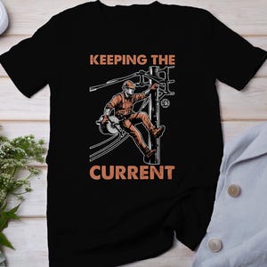 May include: Black t-shirt with a graphic of a lineman working on a power pole. The text "KEEPING THE CURRENT" is printed on the shirt.