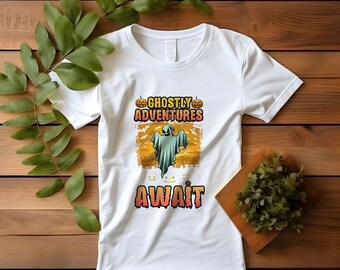May include: White t-shirt with a Halloween graphic. The design features a ghost, pumpkins, and the words "Ghostly Adventures Await" in a colourful, spooky font. The shirt is displayed on a wooden surface with green foliage.