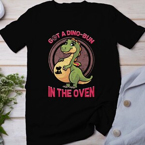 Puede incluir: Camiseta negra con un gráfico de dinosaurio verde. El dinosaurio está embarazada y el texto "Got a Dino-Bun in the Oven" está impreso en la camiseta.