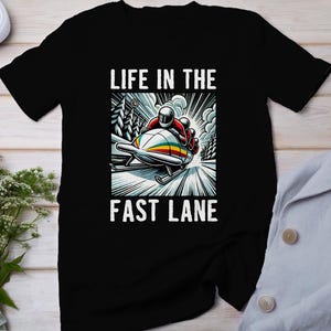 Puede incluir: Camiseta negra con un gráfico de un equipo de bobsleigh corriendo por una pista nevada. El texto "Life in the Fast Lane" está impreso sobre el gráfico.
