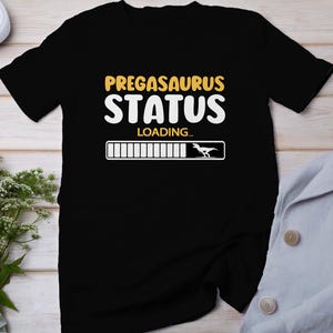 Puede incluir: Camiseta negra con el texto "PREGASAURUS STATUS LOADING..." y una barra de progreso con una silueta de dinosaurio blanca al final.