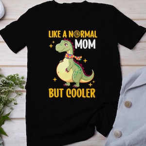 Puede incluir: Camiseta negra con una ilustración de dinosaurio verde y el texto "Como una madre normal pero más cool".
