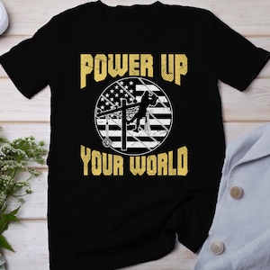 Puede incluir: Camiseta negra con un gráfico amarillo de un lineman trabajando en una línea eléctrica con un diseño de bandera estadounidense. El texto "POWER UP YOUR WORLD" está impreso en amarillo.