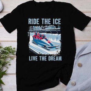 Puede incluir: Camiseta negra con un gráfico de un equipo de bobsleigh corriendo por una pista de hielo. El texto "Ride the Ice" está por encima de la imagen y "Live the Dream" está por debajo.