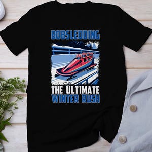 Puede incluir: Camiseta negra con un gráfico de un bobsleigh y el texto "BOBSLEDDING" arriba y "THE ULTIMATE WINTER RUSH" abajo. El diseño incluye un bobsleigh rojo y azul sobre una pista nevada, con un paisaje invernal de fondo.