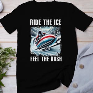 Puede incluir: Camiseta negra con un gráfico de un corredor de bobsleigh en una pista nevada. El texto "Ride the Ice" está por encima del gráfico y "Feel the Rush" está por debajo.