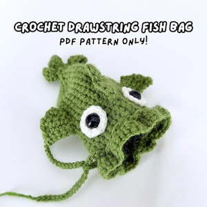 May include: A green crochet drawstring fish bag with black and white plastic eyes and fins. The text CROCHET DRAWSTRING FISH BAG PDF PATTERN ONLY! is printed above the fish-shaped bag, which features a drawstring closure at the open mouth.