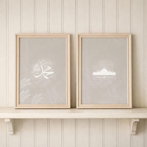 May include: Two framed art prints with light wood frames. One print features white Arabic calligraphy on a gray background. The other print displays a white silhouette of a building on a gray background. The prints are displayed on a white shelf.