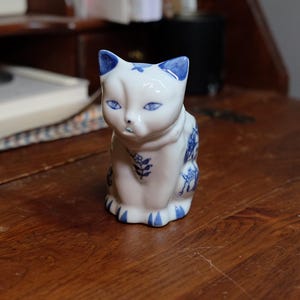 May include: A white ceramic cat figurine with blue accents. The cat has blue eyes, ears, and decorative patterns. The figurine is sitting on a wooden surface.