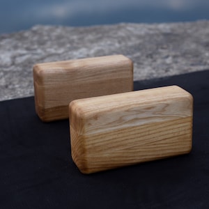 May include: Two rectangular wooden yoga blocks with rounded edges. The blocks are light brown with visible wood grain and are placed on a black mat. The background features a blurred view of water and a stone surface.
