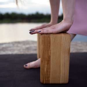 May include: Two rectangular wooden yoga blocks with a natural wood grain are positioned on a black yoga mat. The blocks are being used for support during a yoga pose. The mat has a green border.