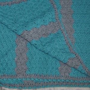 K&ouml;nnte beinhalten: Handgefertigte H&auml;keldecke in T&uuml;rkis und Grau. Die Decke hat ein strukturiertes Stichmuster und einen gewellten Rand. Das Design umfasst eine t&uuml;rkisfarbene Mitte mit grauen, gewellten Bord&uuml;ren und Akzenten. Die Decke ist gefaltet.