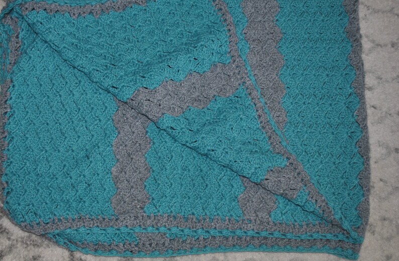 K&ouml;nnte beinhalten: Handgefertigte H&auml;keldecke in T&uuml;rkis und Grau. Die Decke hat ein strukturiertes Stichmuster und einen gewellten Rand. Das Design umfasst eine t&uuml;rkisfarbene Mitte mit grauen, gewellten Bord&uuml;ren und Akzenten. Die Decke ist gefaltet.