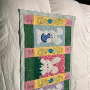 May include: A rectangular Easter-themed fabric wall hanging. It features three panels with embroidered white bunnies holding Easter eggs. The panels are separated by green and yellow sections with decorative stitching and small blue embellishments. The edges are trimmed with a light blue fabric.