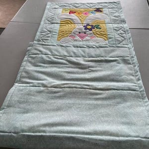 May include: A light blue quilted wall hanging with a bunny design. The bunny has white fur, a pink nose, and a flower on its head. The background is yellow, and the top has a rainbow-colored bow.