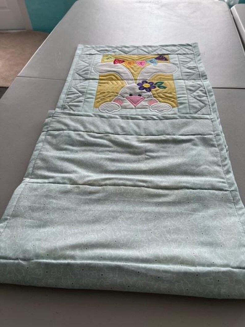 May include: A light blue quilted wall hanging with a bunny design. The bunny has white fur, a pink nose, and a flower on its head. The background is yellow, and the top has a rainbow-colored bow.