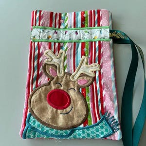 May include: A fabric gift bag featuring a vertical striped pattern in red, white, and green. An embroidered reindeer with a large red nose and a teal patterned scarf is centered on the front. A white band with a string of lights sits above.