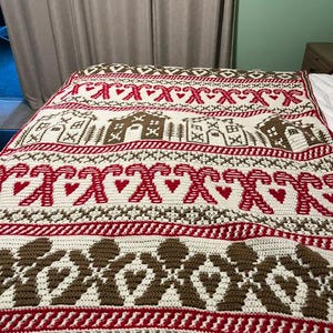 May include: A crocheted blanket featuring a repeating pattern of gingerbread houses, candy canes, hearts, and geometric shapes in red, white, and brown yarn. The textile displays horizontal bands of festive motifs across its entire surface.