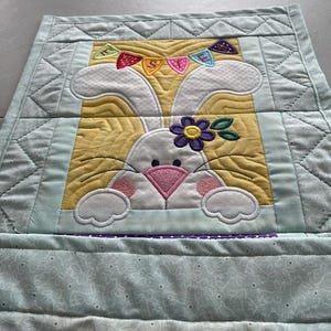 May include: A quilted Easter-themed table runner featuring a white bunny with a pink nose and a purple flower. The bunny is set against a yellow background with a banner that says "EASTER". The runner has a light blue border.