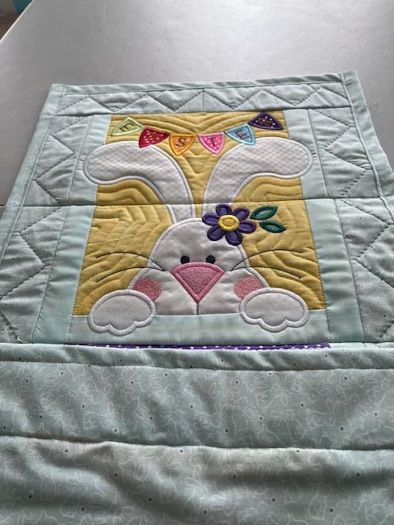 May include: A quilted Easter-themed table runner featuring a white bunny with a pink nose and a purple flower. The bunny is set against a yellow background with a banner that says "EASTER". The runner has a light blue border.