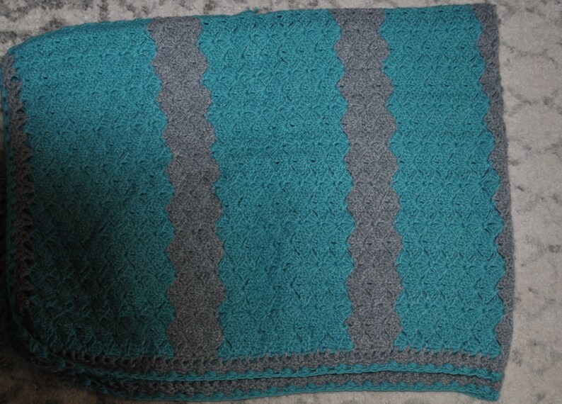 K&ouml;nnte beinhalten: Handgefertigte H&auml;keldecke in T&uuml;rkis und Grau. Die Decke hat abwechselnde vertikale Streifen in T&uuml;rkis und Grau mit einem gewellten Rand. Die Textur der H&auml;kelstiche ist sichtbar und tr&auml;gt zum gem&uuml;tlichen Gef&uuml;hl bei.