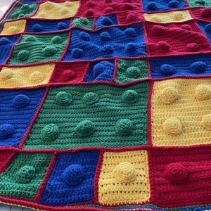 May include: A colorful, handmade crochet blanket with a patchwork design. The blanket features squares of red, blue, green, and yellow, each with textured, raised bobbles. The blanket is edged with a red border.