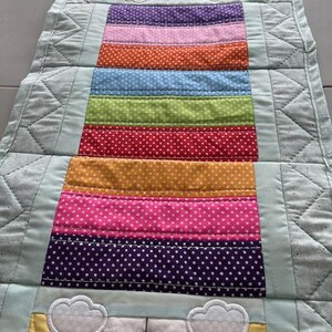 May include: A colorful Easter-themed table runner. The runner features a repeating pattern of a bunny with a flower and a series of horizontal stripes in various colors, including purple, pink, orange, blue, green, red, yellow, and white polka dots. The edges are a light blue.
