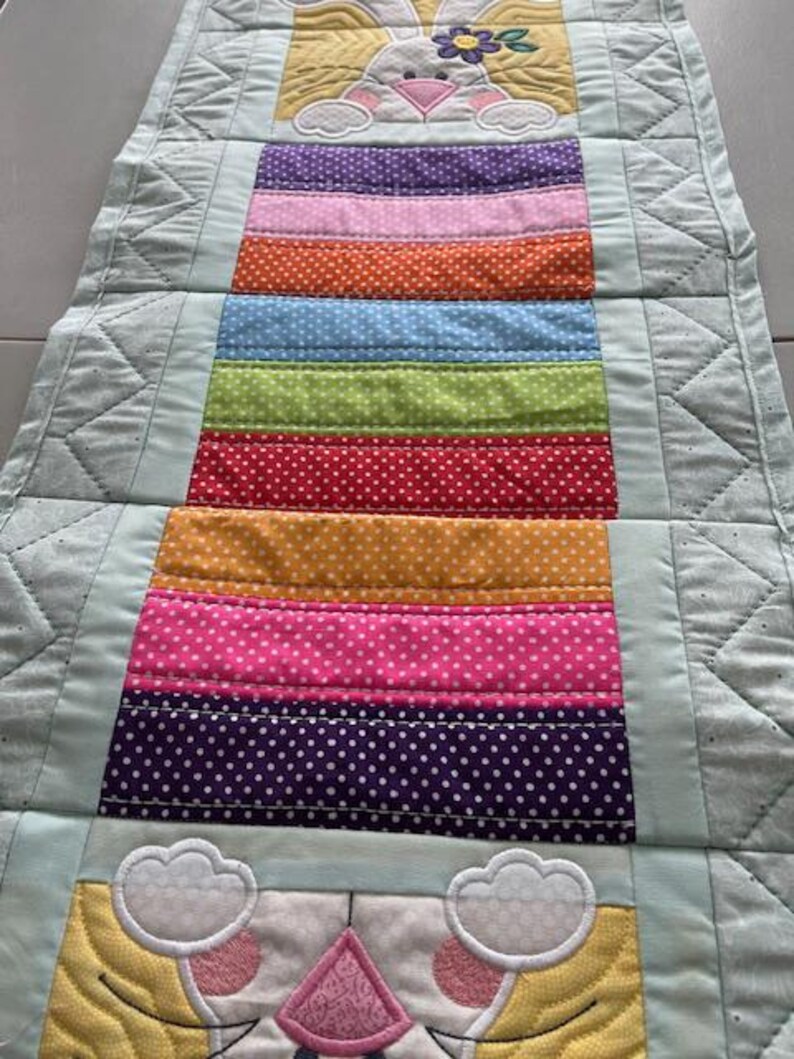 May include: A colorful Easter-themed table runner. The runner features a repeating pattern of a bunny with a flower and a series of horizontal stripes in various colors, including purple, pink, orange, blue, green, red, yellow, and white polka dots. The edges are a light blue.