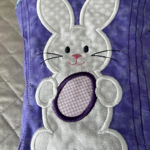 May include: A purple fabric item with a white bunny applique. The bunny has a pink nose, black eyes and whiskers, and is holding a purple and white checkered egg. The bunny is outlined in white stitching.