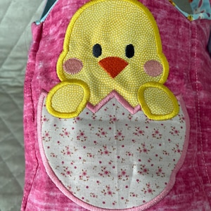 May include: Pink fabric Easter basket with a yellow chick applique. The chick is emerging from a white eggshell with a floral pattern. The basket has a blue handle with a colorful pattern.