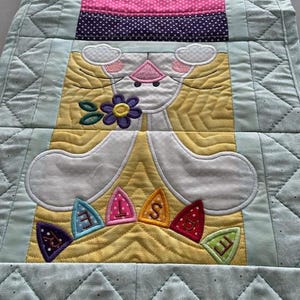 May include: A quilted Easter table runner featuring a bunny design. The bunny is white with pink accents and holds a purple flower. The runner has a light blue border and a striped section with various colors.