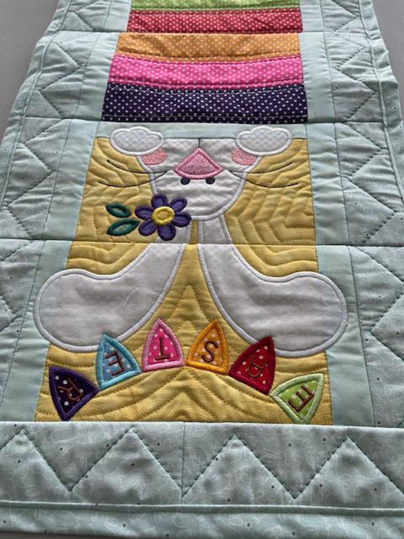 May include: A quilted Easter table runner featuring a bunny design. The bunny is white with pink accents and holds a purple flower. The runner has a light blue border and a striped section with various colors.